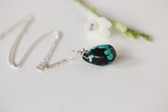 A delicate silver chain necklace with a pendant featuring a black stone with turquoise patterns. The necklace is elegantly laid out on a light surface next to a small white flower with a purple center and green stem.