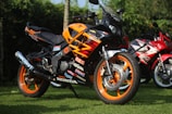 A bright orange and black motorcycle with various decals and branding is parked on a grassy lawn. Another red motorcycle can be seen slightly out of focus in the background, surrounded by green foliage.