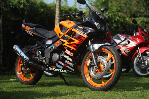 A bright orange and black motorcycle with various decals and branding is parked on a grassy lawn. Another red motorcycle can be seen slightly out of focus in the background, surrounded by green foliage.