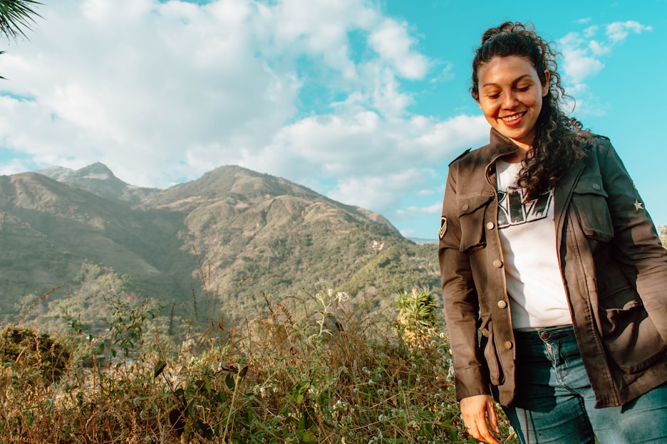 A young Brazilian woman with indigenous features smiling, standing in front of a desert landscape with a mountain of clothes in the background.