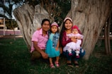 A family of four is sitting in a grassy outdoor area with large tree trunks in the background. The father is wearing a pink shirt, the mother is in a maroon jacket holding a baby in a pink dress, and the young girl in the middle is dressed in a blue outfit and a small tiara.