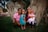 A family of four is sitting in a grassy outdoor area with large tree trunks in the background. The father is wearing a pink shirt, the mother is in a maroon jacket holding a baby in a pink dress, and the young girl in the middle is dressed in a blue outfit and a small tiara.