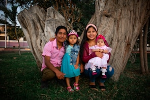 A family of four is sitting in a grassy outdoor area with large tree trunks in the background. The father is wearing a pink shirt, the mother is in a maroon jacket holding a baby in a pink dress, and the young girl in the middle is dressed in a blue outfit and a small tiara.