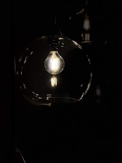 A light bulb emits a warm glow inside a glass enclosure against a dark background. The illumination focuses the viewer's attention on the intricate filaments of the bulb.