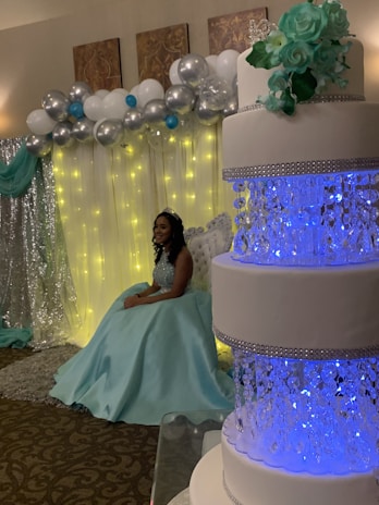A woman in an elegant turquoise gown is sitting beside a large, ornate white cake decorated with blue lighting and floral accents. The background features a decorative wall with silver and white balloons and soft yellow string lights, creating a celebratory atmosphere.