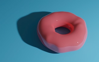 A pink, glossy donut placed on a flat blue surface. The donut casts a soft shadow to the left, emphasizing its three-dimensional form. The lighting creates small white reflections on the donut's glaze, adding to its realistic appearance.