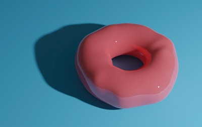A pink, glossy donut placed on a flat blue surface. The donut casts a soft shadow to the left, emphasizing its three-dimensional form. The lighting creates small white reflections on the donut's glaze, adding to its realistic appearance.