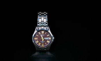 A wristwatch with a metallic strap and a round face with Roman numerals. The watch face is dark with gold-tone hands and markers. The background is solid black, accentuating the timepiece's elegant design.