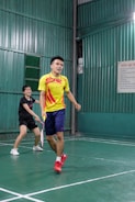 Two people playing badminton in an indoor court, with one person in a yellow shirt and the other in a black shirt. The green walls and flooring create a vibrant backdrop as they focus on their game.