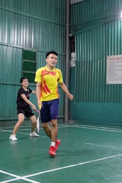 Two people playing badminton in an indoor court, with one person in a yellow shirt and the other in a black shirt. The green walls and flooring create a vibrant backdrop as they focus on their game.