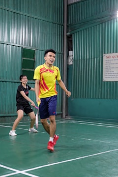 Two people playing badminton in an indoor court, with one person in a yellow shirt and the other in a black shirt. The green walls and flooring create a vibrant backdrop as they focus on their game.