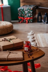 A cozy indoor scene featuring a wooden table with gift boxes wrapped in brown paper and tied with natural twine and ribbon. A small red jar candle is set on the table, surrounded by decorative berries. In the background, larger wrapped gifts are visible, decorated with colorful paper and red ribbons. The setting exudes a warm and festive atmosphere.