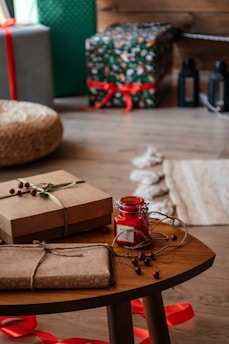A cozy indoor scene featuring a wooden table with gift boxes wrapped in brown paper and tied with natural twine and ribbon. A small red jar candle is set on the table, surrounded by decorative berries. In the background, larger wrapped gifts are visible, decorated with colorful paper and red ribbons. The setting exudes a warm and festive atmosphere.