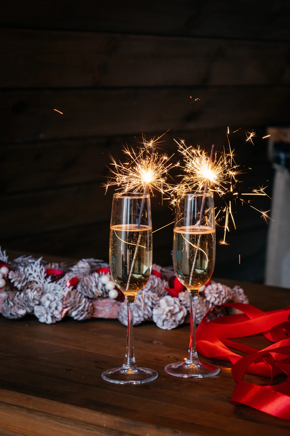 The image shows two champagne flutes filled with a golden liquid, likely champagne, placed on a wooden table draped with a white tablecloth and adorned with red ribbons. The flutes are positioned side by side, each containing a sparkler that is currently lit, creating a warm glow in the scene.
