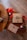 A wooden table displays a neatly wrapped brown gift with a green ribbon and berry decoration, accompanied by red ribbons, jute twine, a patterned envelope, and two jars of scented candles labeled 'Love' and 'Peace'.