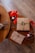 A wooden table displays a neatly wrapped brown gift with a green ribbon and berry decoration, accompanied by red ribbons, jute twine, a patterned envelope, and two jars of scented candles labeled 'Love' and 'Peace'.