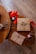 A wooden table displays a neatly wrapped brown gift with a green ribbon and berry decoration, accompanied by red ribbons, jute twine, a patterned envelope, and two jars of scented candles labeled 'Love' and 'Peace'.