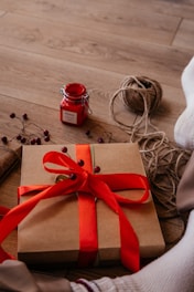 A neatly wrapped gift box with a vibrant red ribbon is placed on wooden flooring. Beside it, there is a small red jar labeled 'peace,' a spool of brown twine, sprigs with small red berries, and part of a person's foot in a sock. The scene suggests a cozy, homemade gift-wrapping session.