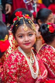 A young girl is dressed elaborately in traditional attire, adorned with ornate jewelry and floral decorations in her hair. She is wearing a richly embroidered red dress with gold accents and layers of pearl necklaces, surrounded by others in a similar festive setting.
