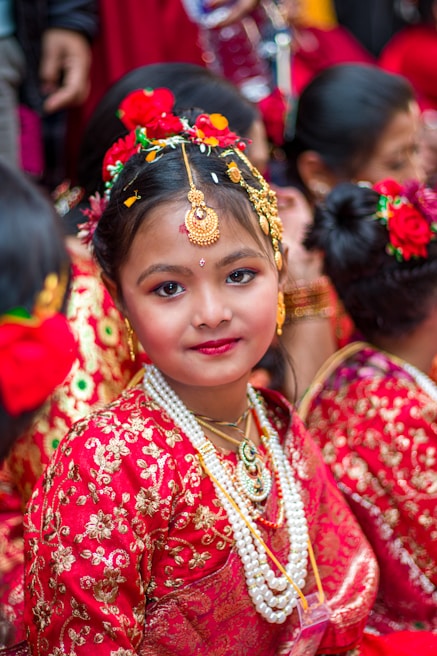 A young girl is dressed elaborately in traditional attire, adorned with ornate jewelry and floral decorations in her hair. She is wearing a richly embroidered red dress with gold accents and layers of pearl necklaces, surrounded by others in a similar festive setting.