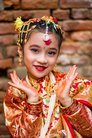 A child dressed in traditional attire with intricate patterns and bright colors. The child has adorned hair with flowers and jewelry, including earrings and a forehead ornament. The vibrant red and gold outfit stands out against the rustic brick background. Her hands are posed gracefully, enhancing the cultural essence of the image.