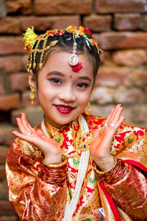 A child dressed in traditional attire with intricate patterns and bright colors. The child has adorned hair with flowers and jewelry, including earrings and a forehead ornament. The vibrant red and gold outfit stands out against the rustic brick background. Her hands are posed gracefully, enhancing the cultural essence of the image.