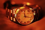 A luxury wristwatch with a metallic strap and a detailed dial featuring Roman numerals. The watch face displays intricate design elements and craftsmanship, with a prominent brand name visible. The overall lighting gives a warm, golden hue to the entire scene, enhancing the watch's elegance.