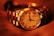 A luxury wristwatch with a metallic strap and a detailed dial featuring Roman numerals. The watch face displays intricate design elements and craftsmanship, with a prominent brand name visible. The overall lighting gives a warm, golden hue to the entire scene, enhancing the watch's elegance.