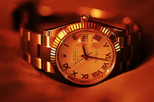 A luxury wristwatch with a metallic strap and a detailed dial featuring Roman numerals. The watch face displays intricate design elements and craftsmanship, with a prominent brand name visible. The overall lighting gives a warm, golden hue to the entire scene, enhancing the watch's elegance.