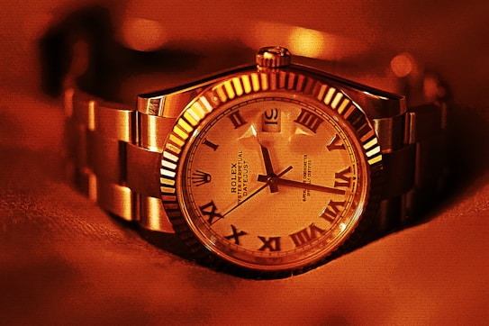 A luxury wristwatch with a metallic strap and a detailed dial featuring Roman numerals. The watch face displays intricate design elements and craftsmanship, with a prominent brand name visible. The overall lighting gives a warm, golden hue to the entire scene, enhancing the watch's elegance.