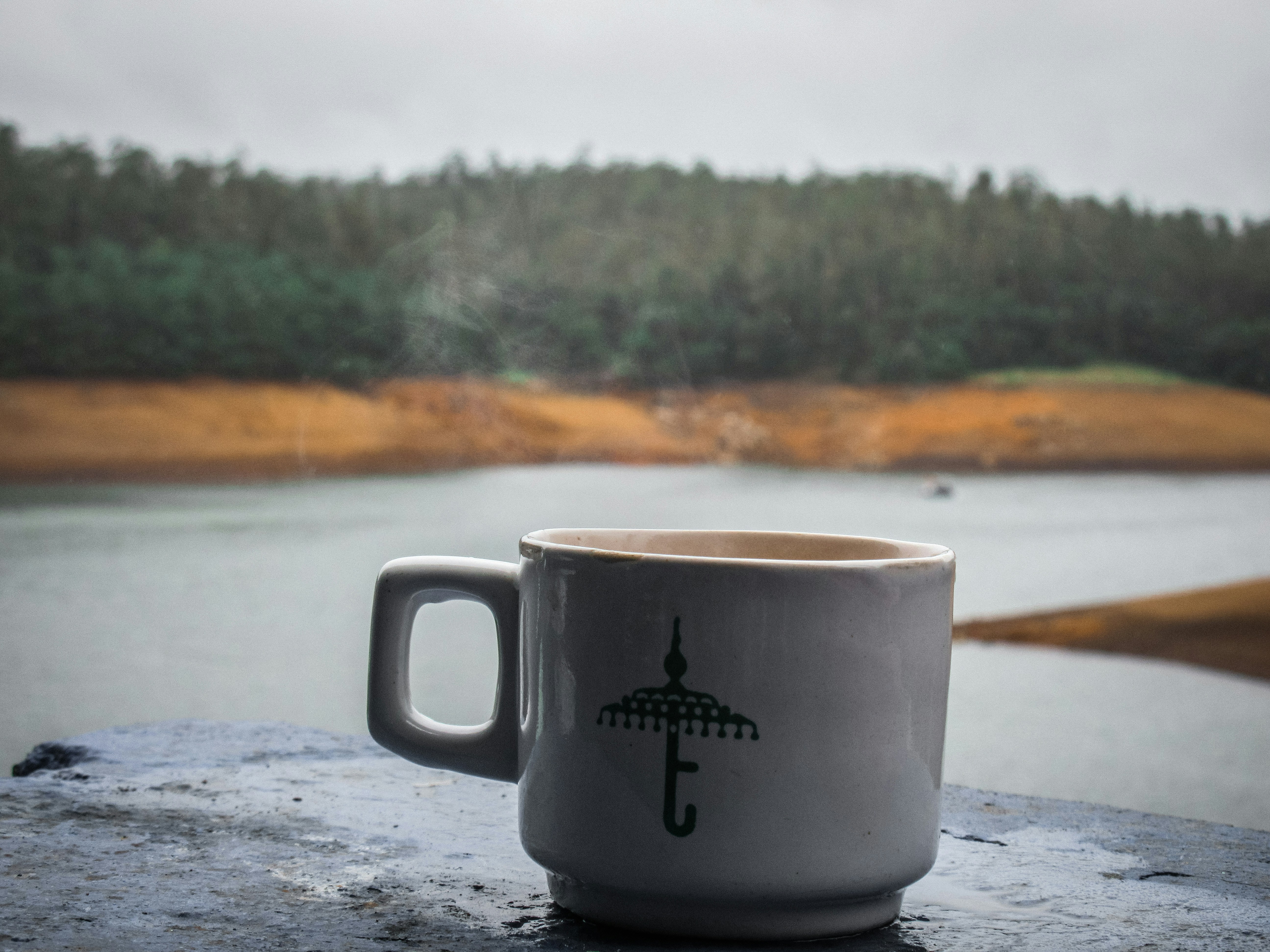 Ceramic mug with an umbrella logo sits on a weathered surface in the foreground, while a calm river and forested shoreline recede in soft distance.