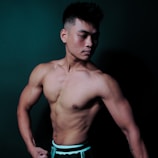 A muscular man with a fit physique poses confidently against a dark background. His expression is serious, and he is shirtless, showcasing his toned torso and arms. The lighting emphasizes his muscle definition and contours.