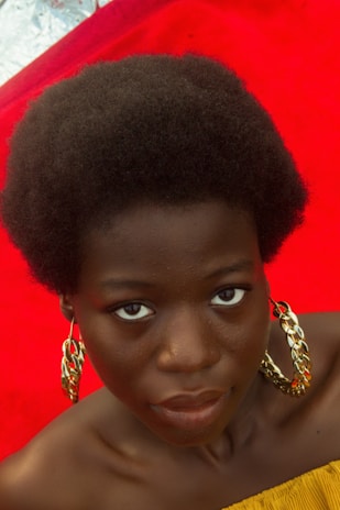 A person with short curly hair and large golden hoop earrings gazes directly at the camera. They are wearing a strapless yellow top. The background features a vibrant red carpet.