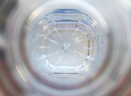 The image captures the view from the bottom inside a clear plastic bottle. The pattern of the bottle’s base features multiple ridges radiating outward from the center, which is a small circular indentation. The plastic is slightly reflective, giving off a translucent, almost glass-like appearance.