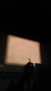 A dark room with a light beige rectangular projection on the wall. A shadow of a hand holding a pen or stylus is visible, suggesting writing or drawing. The lower part of the wall has a border with heart shapes.