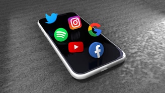A smartphone lies on a textured gray surface with three-dimensional logos of popular apps and platforms, including Twitter, Instagram, Google, Spotify, YouTube, and Facebook, seemingly floating above the screen.