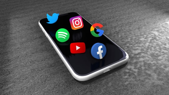 A smartphone lies on a textured gray surface with three-dimensional logos of popular apps and platforms, including Twitter, Instagram, Google, Spotify, YouTube, and Facebook, seemingly floating above the screen.