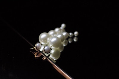 A collection of spherical, white pearls of varying sizes is arranged on a reflective surface. The pearls appear to form a cluster, with light subtly highlighting their smooth, shiny texture against a dark background.