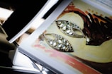 A pair of ornate, teardrop-shaped earrings with multiple gemstones are laid on top of an open book showing a colorful painting. The earrings have a delicate design with white and clear stones set in a gold frame. The painting underneath features abstract shapes and a palette of earth tones including brown, red, and beige.