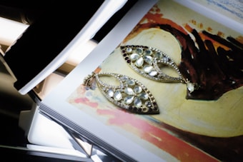 A pair of ornate, teardrop-shaped earrings with multiple gemstones are laid on top of an open book showing a colorful painting. The earrings have a delicate design with white and clear stones set in a gold frame. The painting underneath features abstract shapes and a palette of earth tones including brown, red, and beige.
