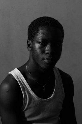 A man with short curly hair gazes directly at the camera against a plain backdrop. The lighting is low-key, casting deep shadows on the right side of his face, while a single light source highlights the left side. He is wearing a sleeveless white shirt and a chain necklace with a cross pendant.