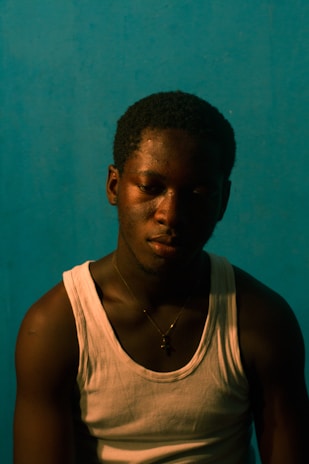 A young man with a somber expression is wearing a white tank top and a necklace with a cross pendant. The background is a plain teal color, and the lighting is dim, casting shadows on his face.