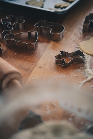 Various metal cookie cutters are scattered on a wooden surface near a rolling pin and some rolled-out dough sprinkled with flour. A baking tray with partially cut-out cookies is visible in the background.