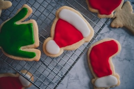 A selection of festive cookies decorated with vibrant icing, including a red Santa hat, a green Christmas tree, and other holiday-themed designs, placed on a cooling rack. The cookies are set against a marble surface, enhancing the festive mood with their bright, colorful icing.