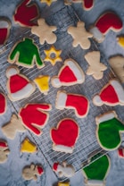 A variety of festive cookies shaped like holiday-themed items rest on a cooling rack. The cookies are decorated with bright red, green, yellow, and white icing, depicting shapes such as mittens, Santa hats, stockings, Christmas sweaters, stars, and gingerbread people. The background is a marble surface, adding a sophisticated touch to the arrangement.