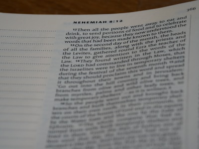 A close-up view of a page from a book, specifically from the book of Nehemiah 8:12. The text is slightly blurred towards the bottom, with clear headings and verses that discuss topics related to food, celebration, and religious events. The page includes a column of dotted lines on the left.