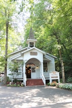 A quaint chapel is nestled among lush green trees. The small, white-painted building features a sharp steeple, a welcoming front porch with steps, and decorative plants lining the entry. Sunlight filters through the leaves, creating a peaceful and serene ambiance.
