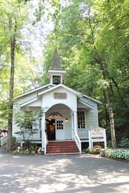 A quaint chapel is nestled among lush green trees. The small, white-painted building features a sharp steeple, a welcoming front porch with steps, and decorative plants lining the entry. Sunlight filters through the leaves, creating a peaceful and serene ambiance.