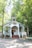 A quaint chapel is nestled among lush green trees. The small, white-painted building features a sharp steeple, a welcoming front porch with steps, and decorative plants lining the entry. Sunlight filters through the leaves, creating a peaceful and serene ambiance.