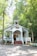 A quaint chapel is nestled among lush green trees. The small, white-painted building features a sharp steeple, a welcoming front porch with steps, and decorative plants lining the entry. Sunlight filters through the leaves, creating a peaceful and serene ambiance.